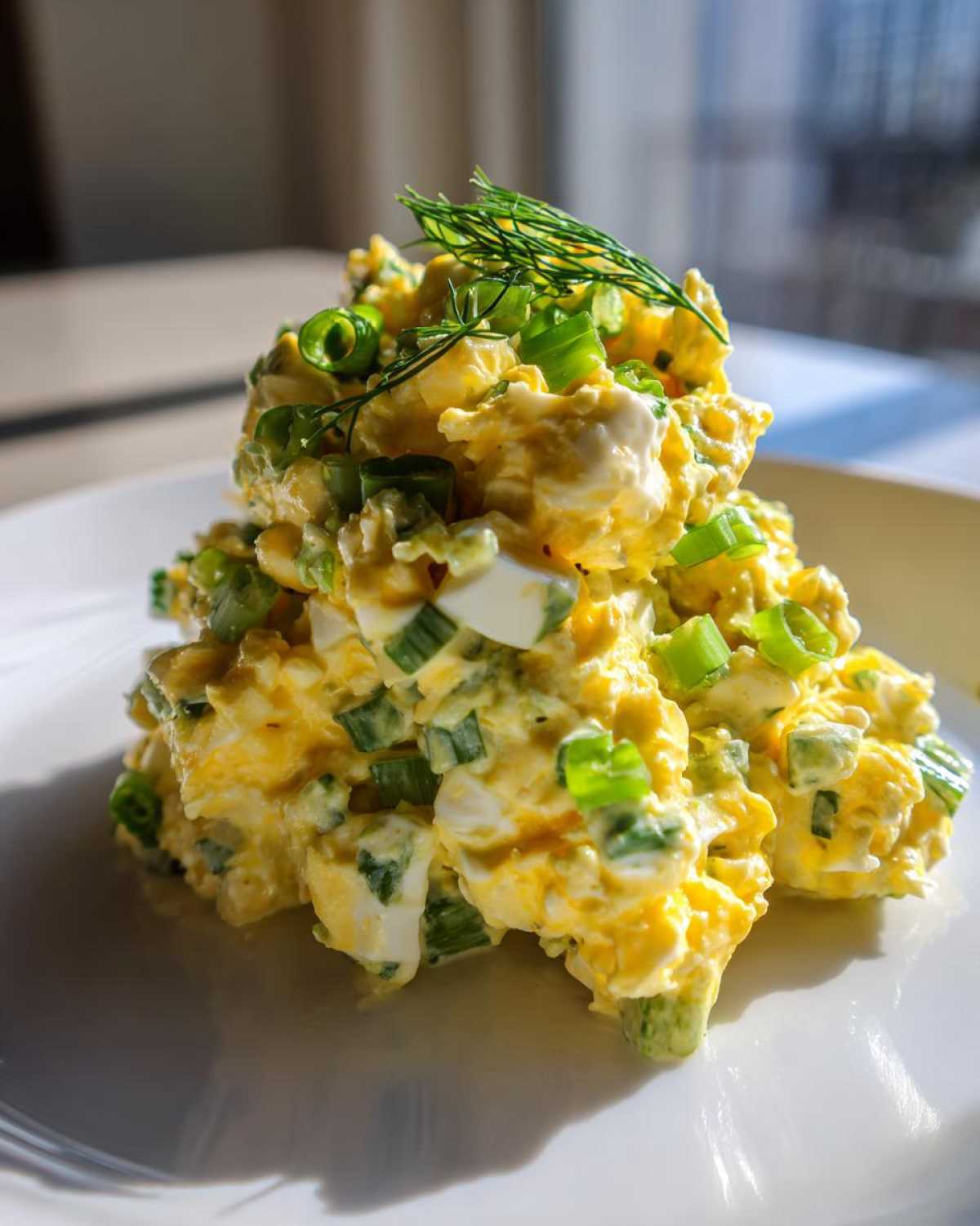 A mound of creamy egg salad recipe mixed with green onions and topped with fresh dill on a white plate.