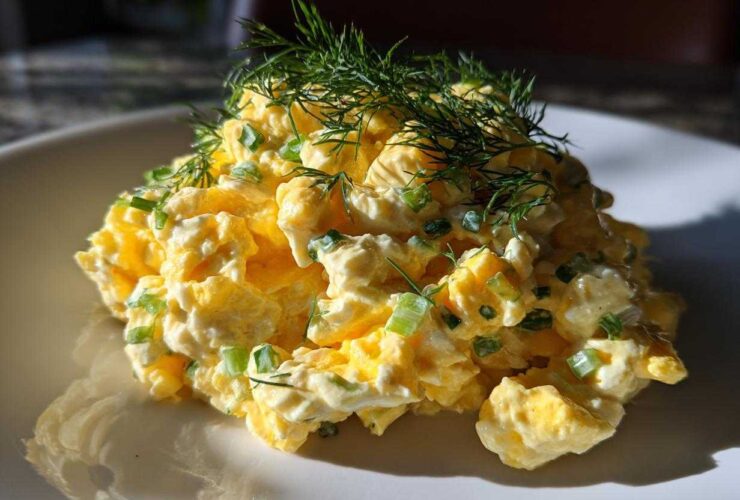 A mound of creamy egg salad recipe garnished with chopped green onions and fresh dill sprigs on a white plate.
