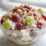 A close-up of creamy grape salad, featuring red and green grapes mixed with a sweet cream dressing and topped with chopped pecans.