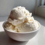 Three generous scoops of creamy, white protein ice cream served in a bright white bowl, catching natural sunlight.