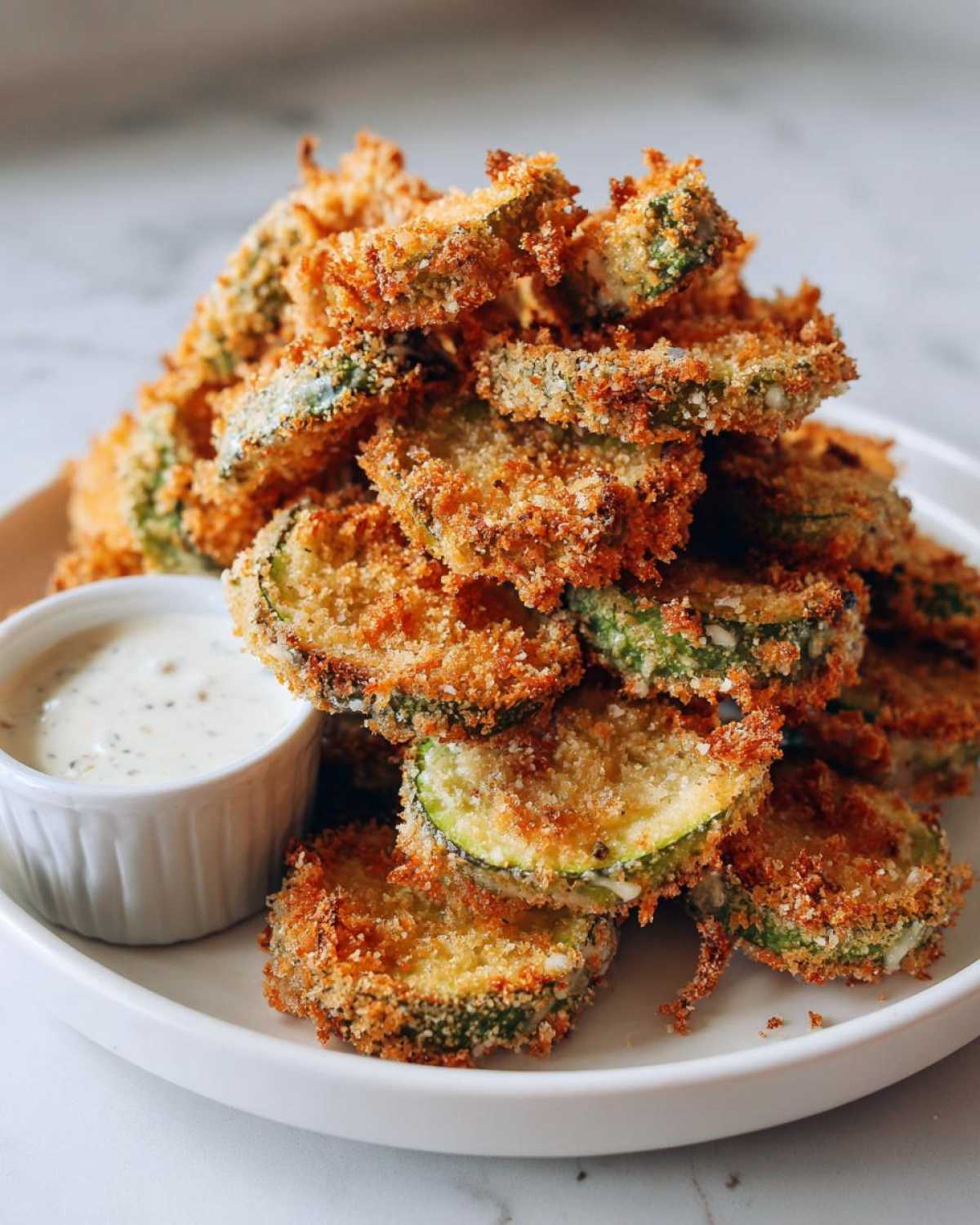 A tall stack of golden brown, crispy Fried Pickles served on a white plate next to a small ramekin of creamy dipping sauce.