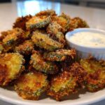 A generous pile of golden brown, crispy fried pickles served on a white plate next to a small bowl of creamy dipping sauce.