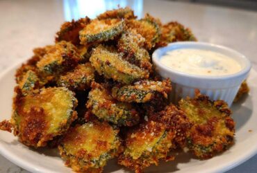 A generous pile of golden brown, crispy fried pickles served on a white plate next to a small bowl of creamy dipping sauce.