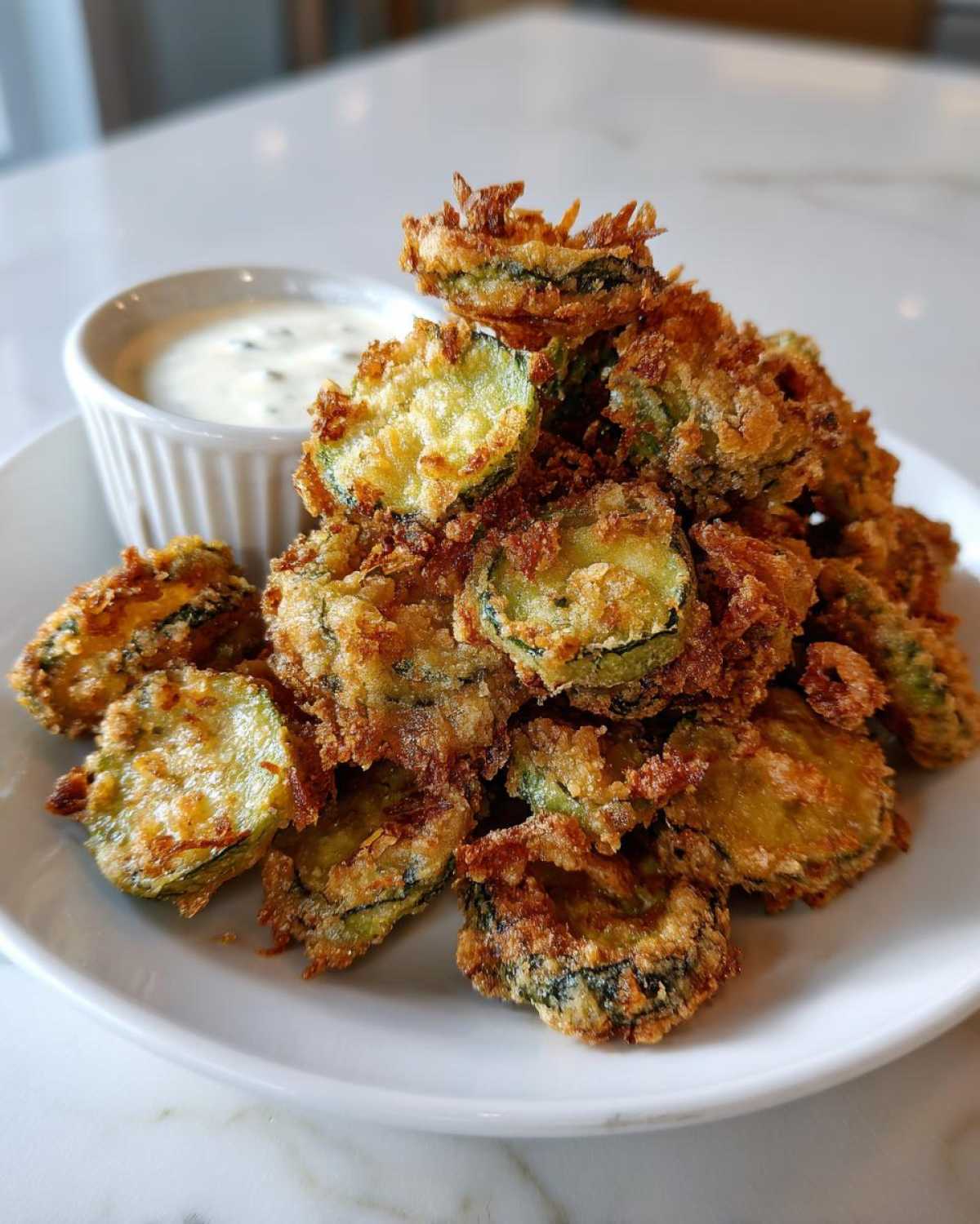 A large pile of golden brown, crispy Fried Pickles served on a white plate next to a small ramekin of creamy dipping sauce.