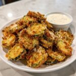 A mound of golden brown, crispy fried pickles served on a white plate next to a small bowl of creamy dipping sauce.