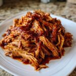A close-up of shredded, tender crock pot pulled pork generously coated in rich, dark barbecue sauce on a white plate.