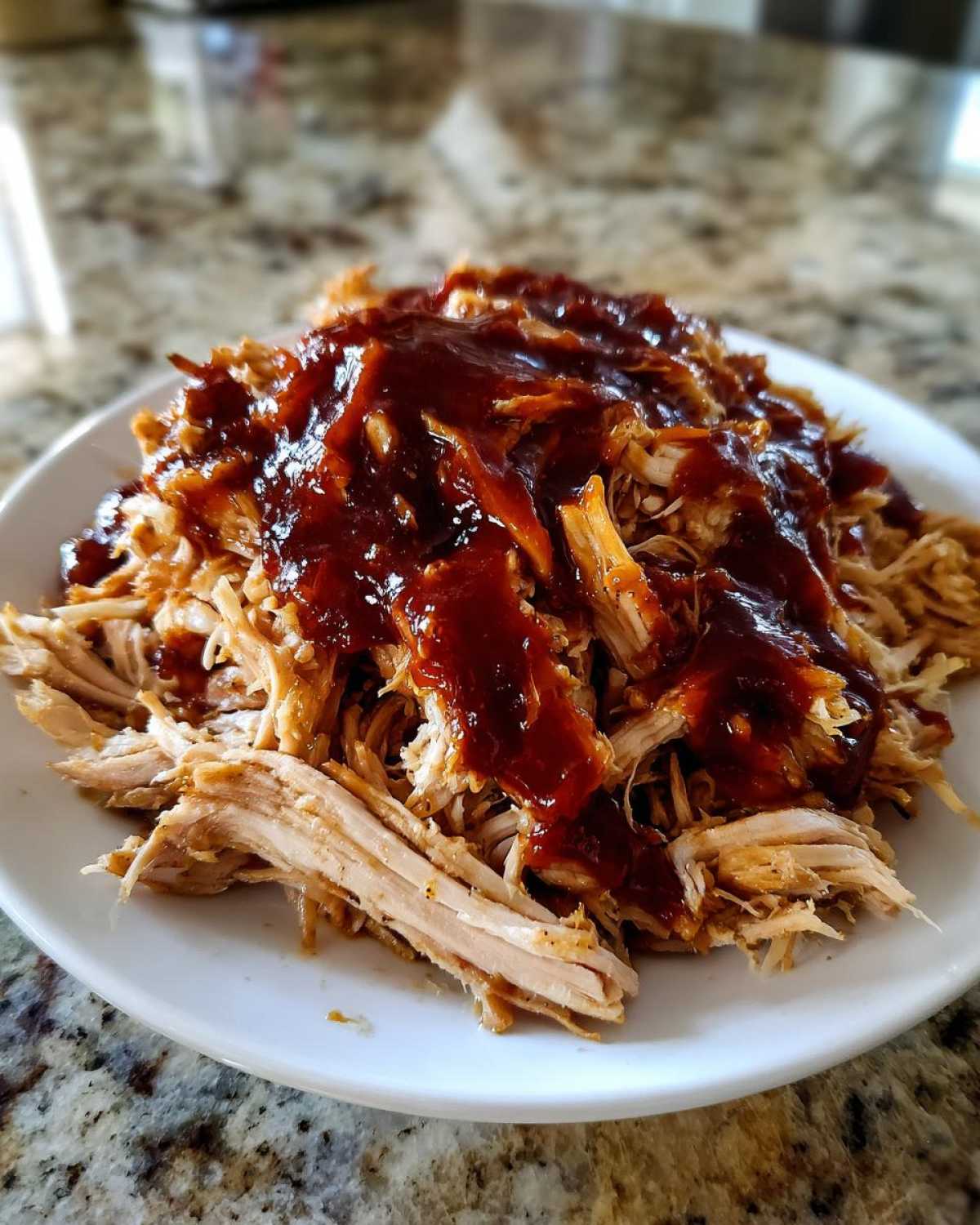 Close-up of shredded crock pot pulled pork generously topped with rich, dark BBQ sauce on a white plate.