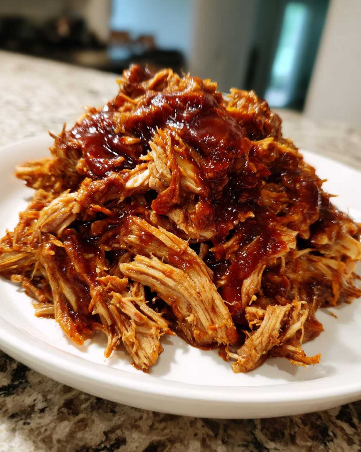A close-up of a generous mound of shredded crock pot pulled pork drizzled with thick barbecue sauce, served on a white plate.
