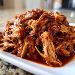 A close-up of shredded, saucy crock pot pulled pork piled high on a white rectangular plate.