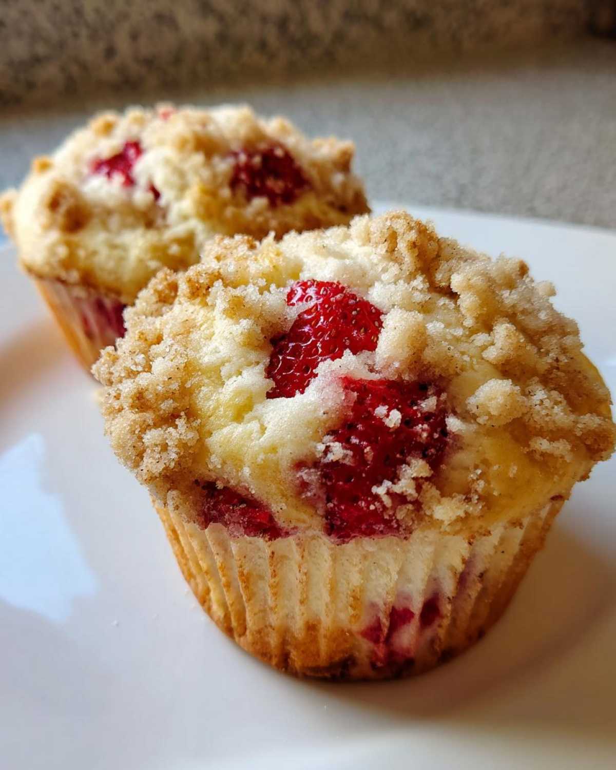 Two delicious strawberry muffins with a sweet crumb topping resting on a white plate.