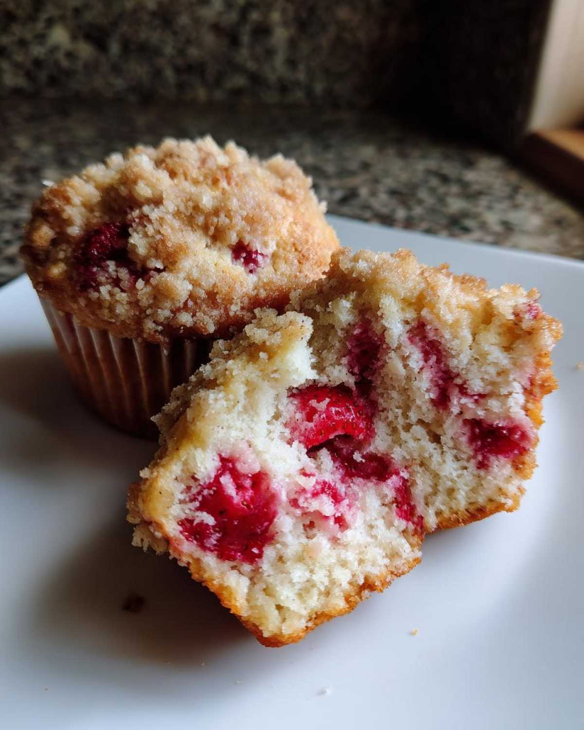 A close-up of a crumb-topped strawberry muffin cut in half, showing the moist interior packed with bright red strawberries.