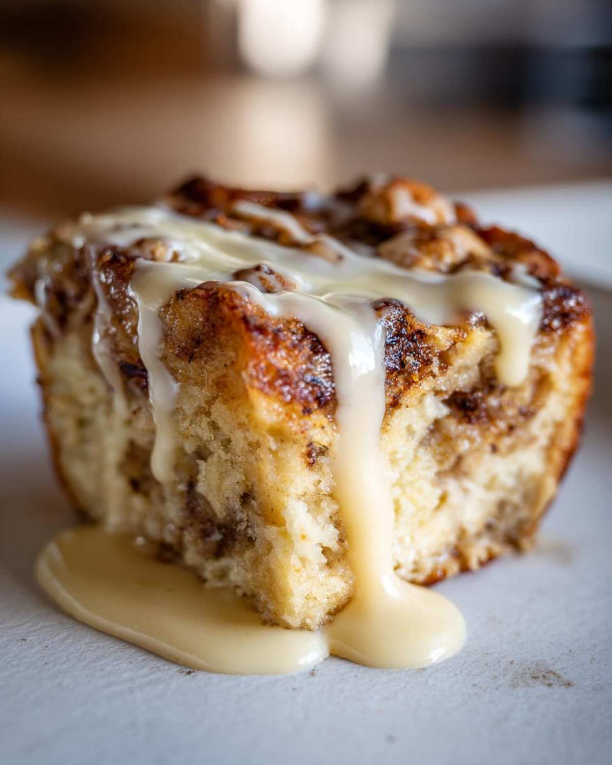 A close-up shot of a serving of rich bread pudding topped with thick, creamy vanilla glaze dripping down the side.