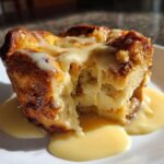 A close-up of a portion of warm bread pudding, drizzled heavily with a creamy vanilla sauce, served on a white plate.