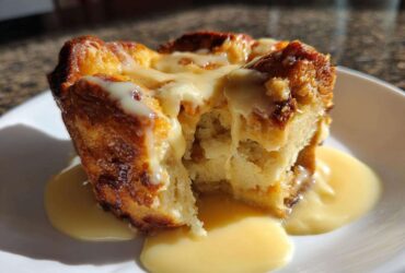 A close-up of a portion of warm bread pudding, drizzled heavily with a creamy vanilla sauce, served on a white plate.