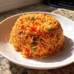 A molded serving of vibrant, orange-red spanish rice mixed with peas and diced tomatoes in a white bowl.
