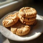 A stack and pile of freshly baked Easy Peanut Butter Cookies with classic crisscross fork marks on a white plate.