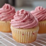 Three delicious strawberry cupcakes with light yellow cake bases and vibrant pink, swirled frosting speckled with berry pieces.