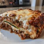 A close-up of a hearty slice of zucchini lasagna, showing layers of meat sauce, cheese, and a visible zucchini noodle.
