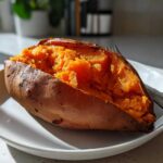 A close-up of a perfectly baked sweet potato split open, showing its bright orange, fluffy interior, resting on a white plate.