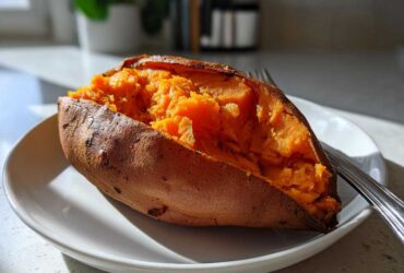 A close-up of a perfectly baked sweet potato split open, showing its bright orange, fluffy interior, resting on a white plate.