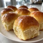 A close-up of several golden-brown, fluffy dinner rolls glistening with melted butter on a white plate.