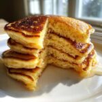 Close-up of a tall stack of fluffy greek yogurt pancakes with a piece cut out, drizzled with syrup.