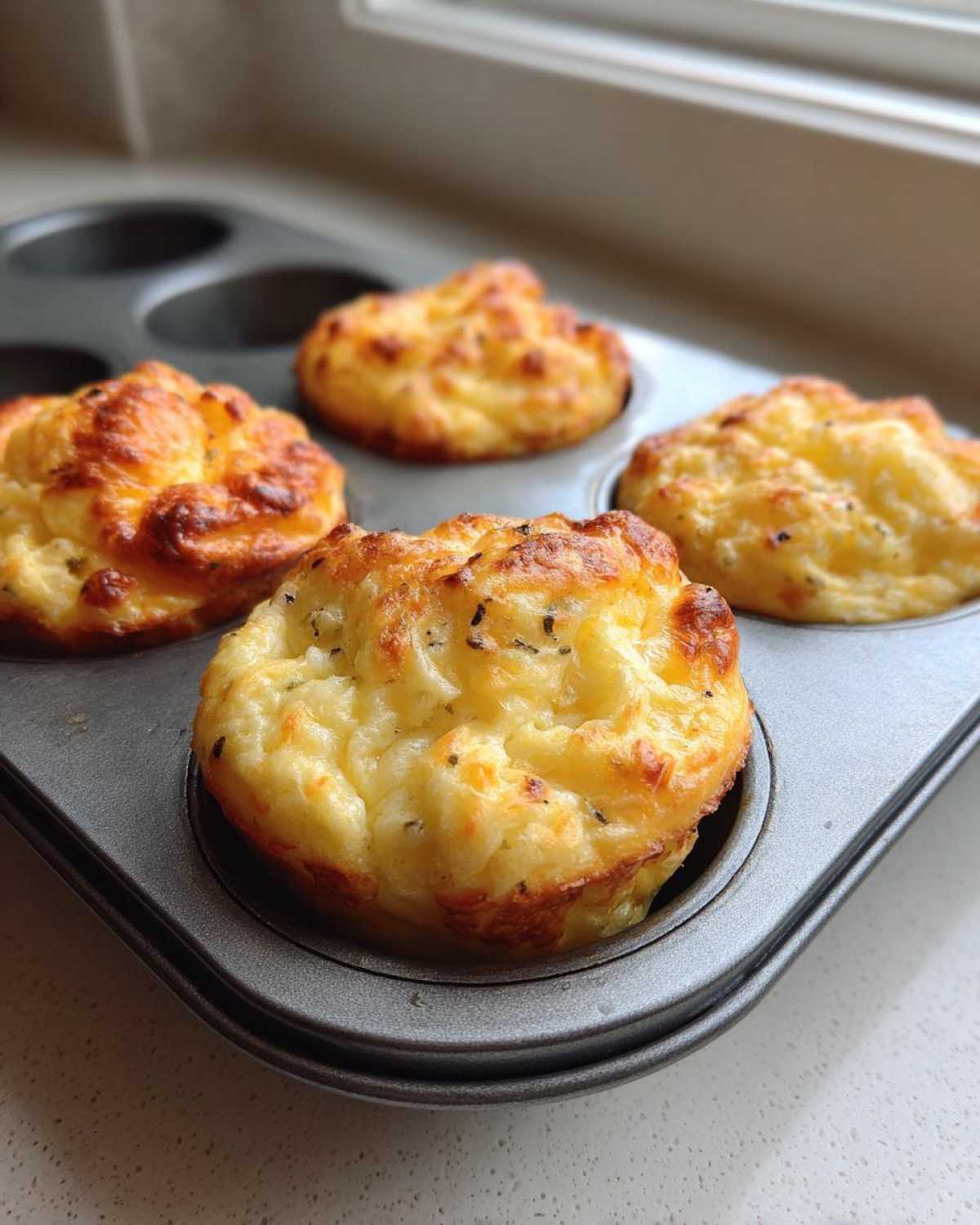 Four golden brown, fluffy muffin tin egg bites resting in a dark grey non-stick muffin pan.