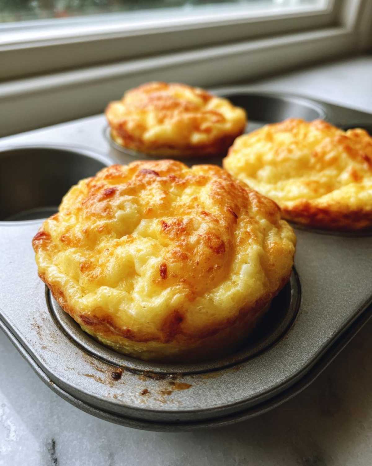Close-up of three fluffy muffin tin egg bites with golden-brown tops resting in a metal muffin pan.