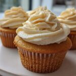 A close-up photograph of several moist vanilla cupcakes topped with a generous swirl of light cream-colored frosting.