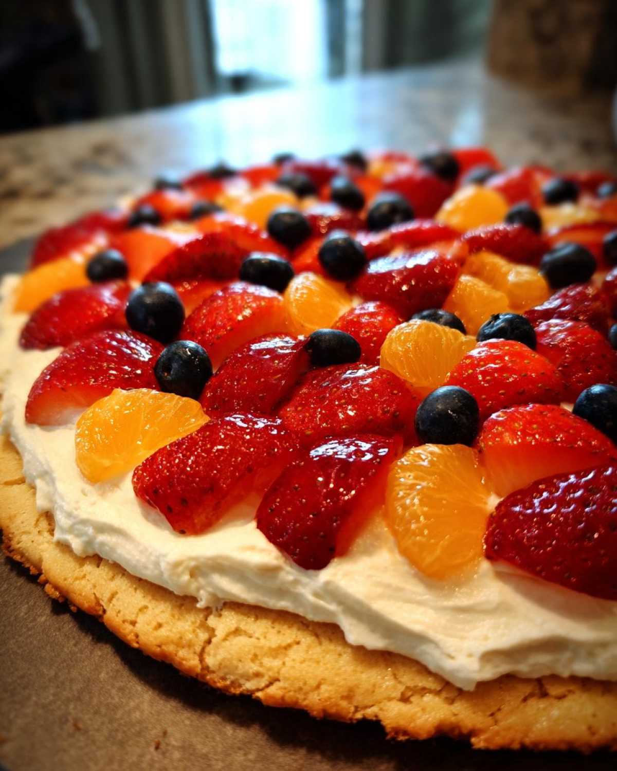 A close-up view of a fruit pizza sugar cookie topped with whipped cream, strawberries, blueberries, and mandarin oranges.