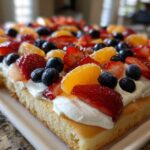 Close-up of a rectangular fruit pizza sugar cookie topped with whipped cream, strawberries, blueberries, and mandarin oranges.