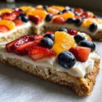 Close-up of a slice of fruit pizza sugar cookie topped with cream cheese frosting, strawberries, blueberries, and orange segments.