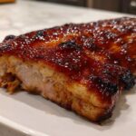 Close-up of a piece of meat, likely BBQ ribs, covered in a thick, glossy, dark brown glaze, served on a white plate.
