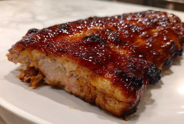 Close-up of a piece of meat, likely BBQ ribs, covered in a thick, glossy, dark brown glaze, served on a white plate.