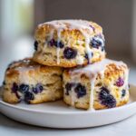 Three fluffy blueberry scones stacked on a white plate, generously topped with a sweet white glaze.