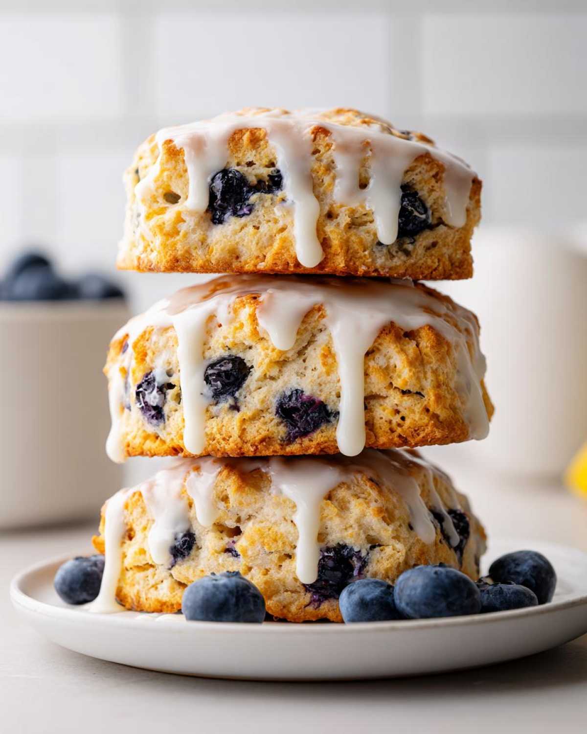 Three delicious blueberry scones stacked vertically, topped with white glaze, resting on a plate with fresh blueberries.