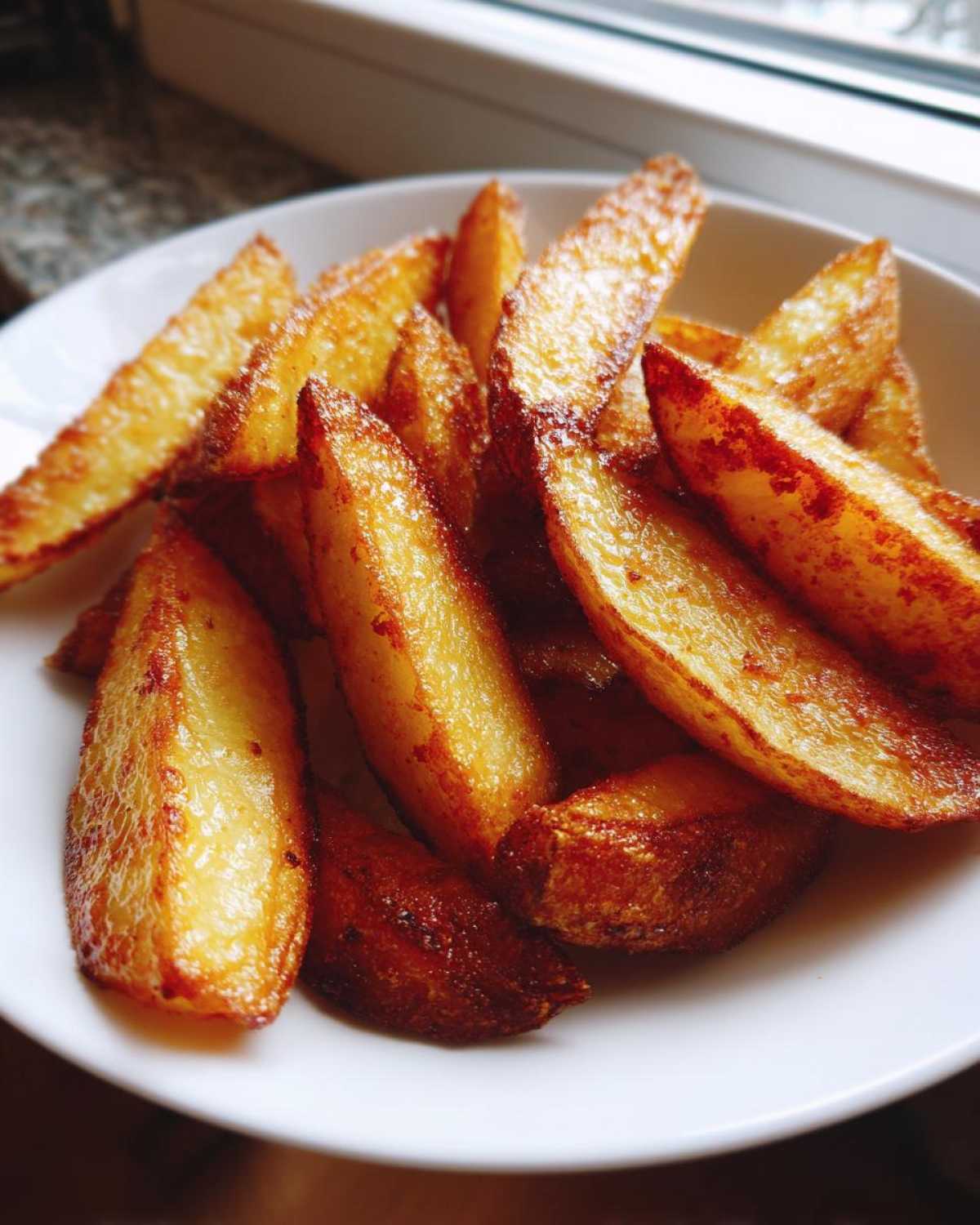 Close-up of perfectly cooked, golden brown air fryer potato wedges piled high on a white dish near a window.