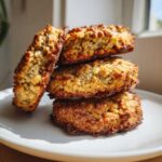 A stack of four golden brown, textured breakfast cookie treats resting on a white plate near a bright window.