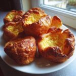Several golden brown, deeply risen yorkshire pudding towers resting on a white plate near a window.