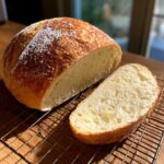 A freshly baked loaf of italian bread, partially sliced, showing the soft interior and golden crust, resting on a cooling rack.