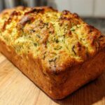 A freshly baked, golden brown loaf of Italian herb and cheese bread resting on a wooden cutting board.