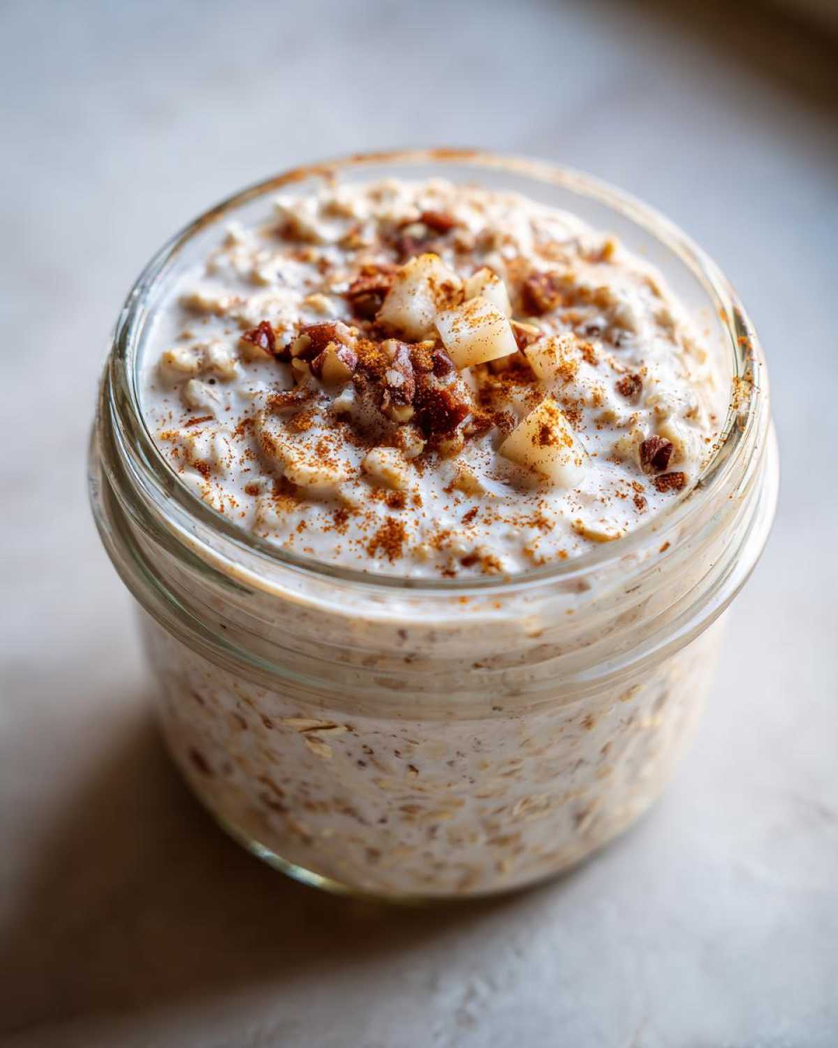 Close-up of apple cinnamon overnight oats in a glass jar, topped with diced apple, pecans, and cinnamon.