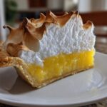 A close-up of a tall slice of lemon meringue pie showing bright yellow filling and peaks of browned meringue.