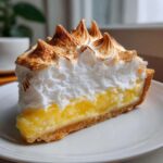 A close-up of a tall slice of lemon meringue pie showing bright yellow filling and fluffy, torched meringue peaks.