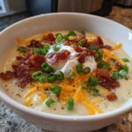 A close-up of a bowl of creamy loaded baked potato soup topped with sour cream, shredded cheddar, bacon bits, and green onions.