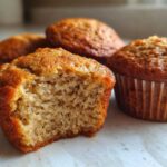 Close-up of a banana protein muffin with a bite taken out, showing the moist, textured interior next to whole muffins.