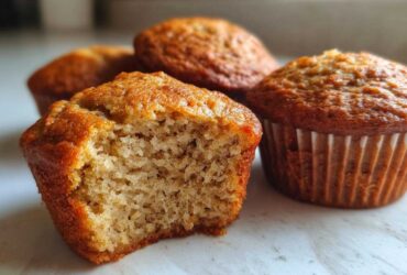 Close-up of a banana protein muffin with a bite taken out, showing the moist, textured interior next to whole muffins.