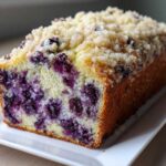 A close-up of a moist blueberry banana bread loaf, showing the dense crumb packed with blueberries and topped with a sugary streusel.