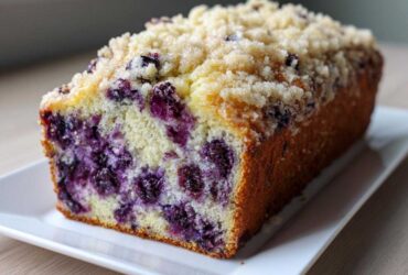 A close-up of a moist blueberry banana bread loaf, showing the dense crumb packed with blueberries and topped with a sugary streusel.