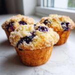 Three moist blueberry muffins topped with a crunchy sugar crumb topping sitting on a white marble surface.
